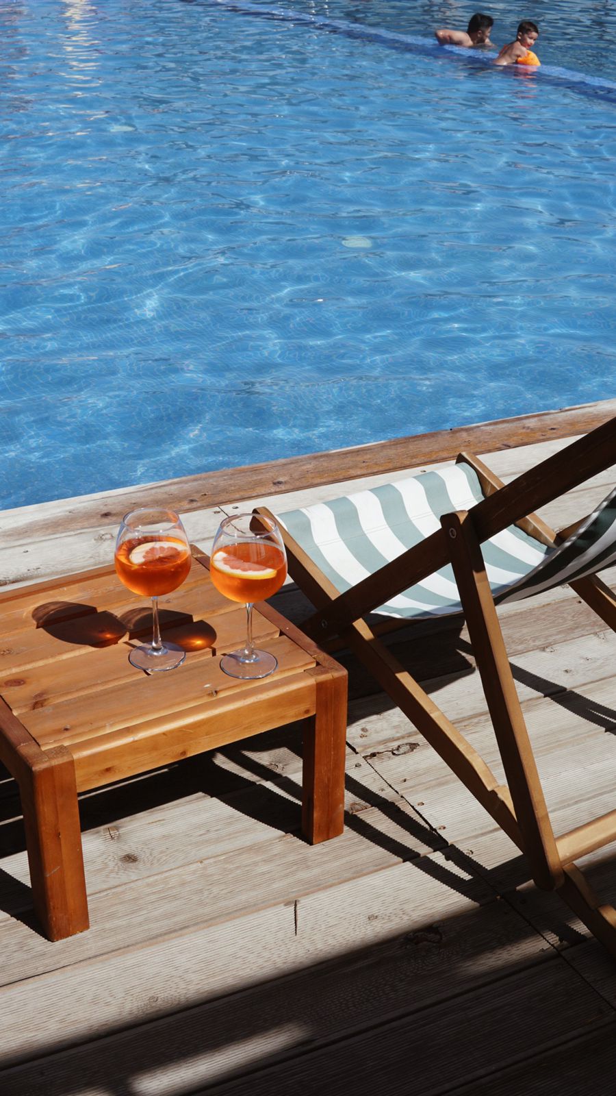 Two glasses filled with orange beverages are placed on a wooden side table beside a striped lounge chair. A clear blue swimming pool reflects sunlight, with two individuals swimming in the background.