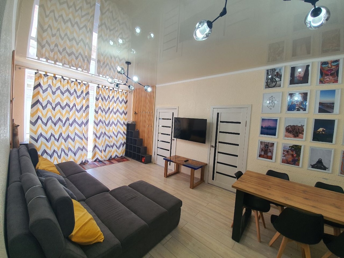A spacious living area is showcased with a large, dark grey sectional sofa adorned with yellow cushions. A smart television is mounted on the wall opposite the sofa. Natural light enters through patterned curtains, illuminating wooden accents and a dining table with black chairs.