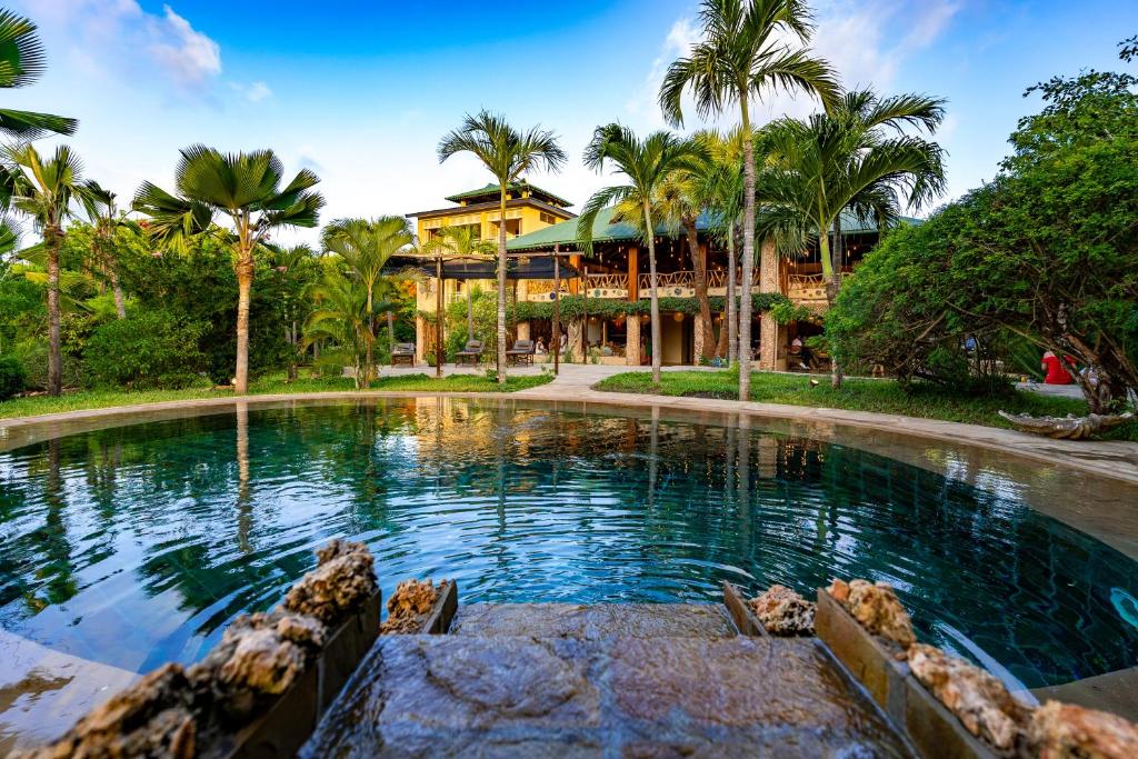 A tranquil pool is framed by lush palm trees, reflecting the surrounding greenery. The serene water features gentle ripples and is bordered by natural stone. In the background, a multi-level structure with an open terrace is visible, enhancing the peaceful ambiance of the outdoor space.