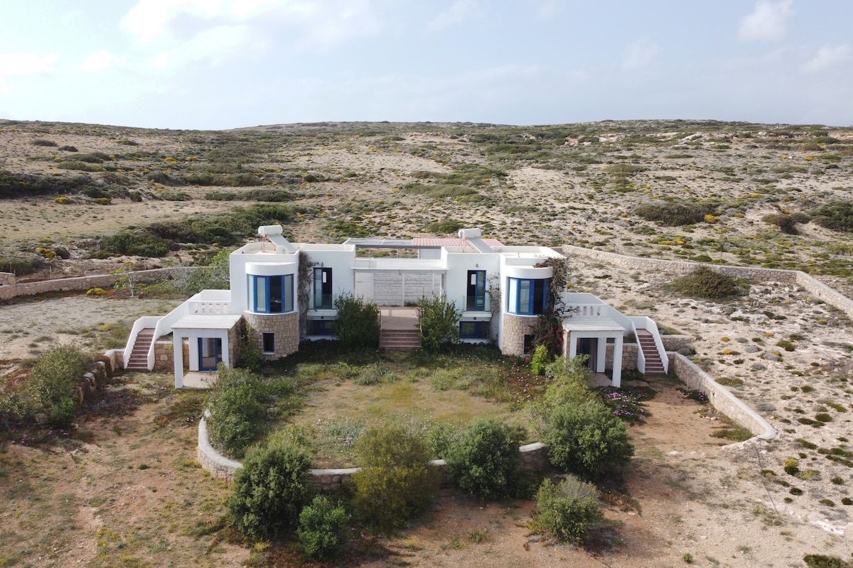 The house is showcased from an aerial view, featuring a unique architectural design with rounded structures. Surrounding greenery and low-lying plants are visible, along with a natural landscape that extends to the horizon. The perimeter is defined by a low stone wall.