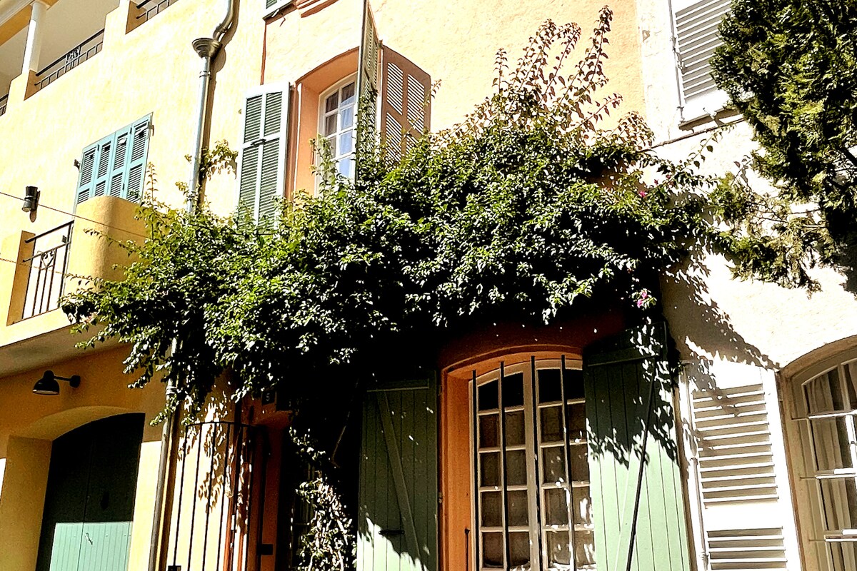 The exterior of the house features a warm yellow façade adorned with lush greenery, including climbing plants that enhance the entrance. Large wooden doors in soft green and white shutters frame the windows, inviting a serene and charming atmosphere.