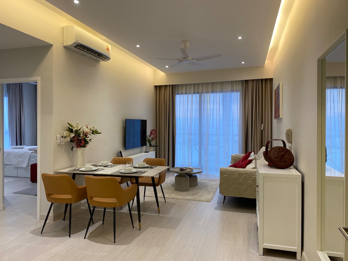 A modern living area features a dining table with four chairs, accompanied by a cozy sofa beside a large window. A stylish television is mounted on the wall, while soft lighting enhances the open and airy ambiance. Decorative elements include flowers and artwork.