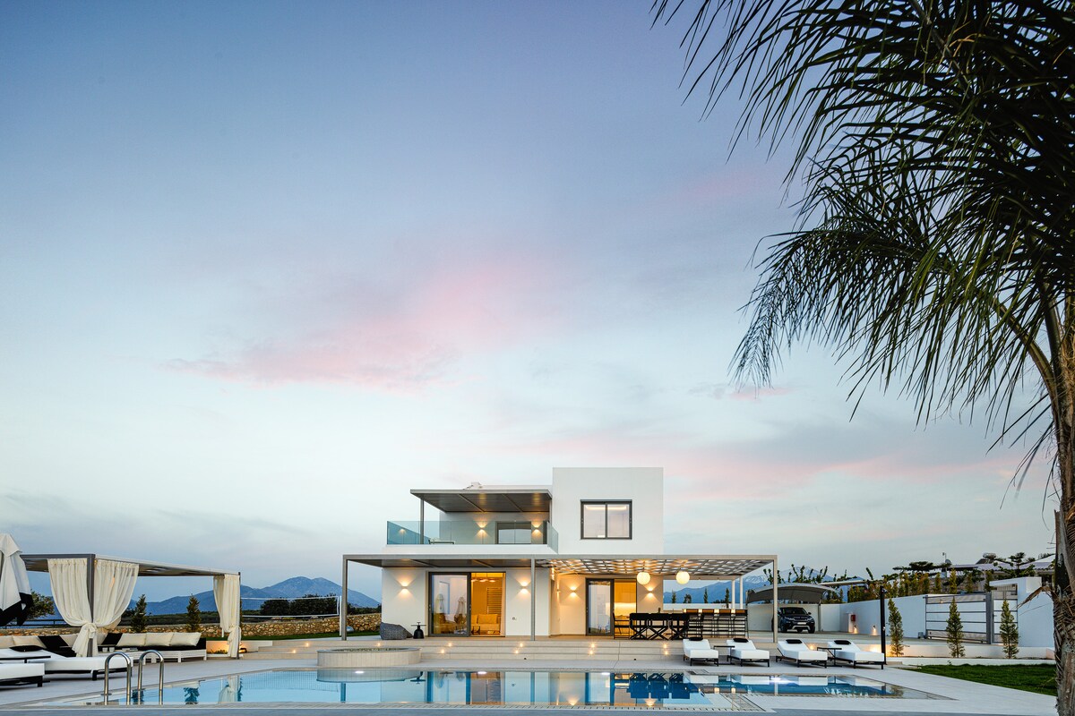 A modern villa is set against a soft twilight sky, highlighted by gentle hues of pink and blue. The structure features expansive windows and a spacious terrace overlooking the private pool, complemented by stylish sun loungers and shaded seating areas amidst lush greenery.