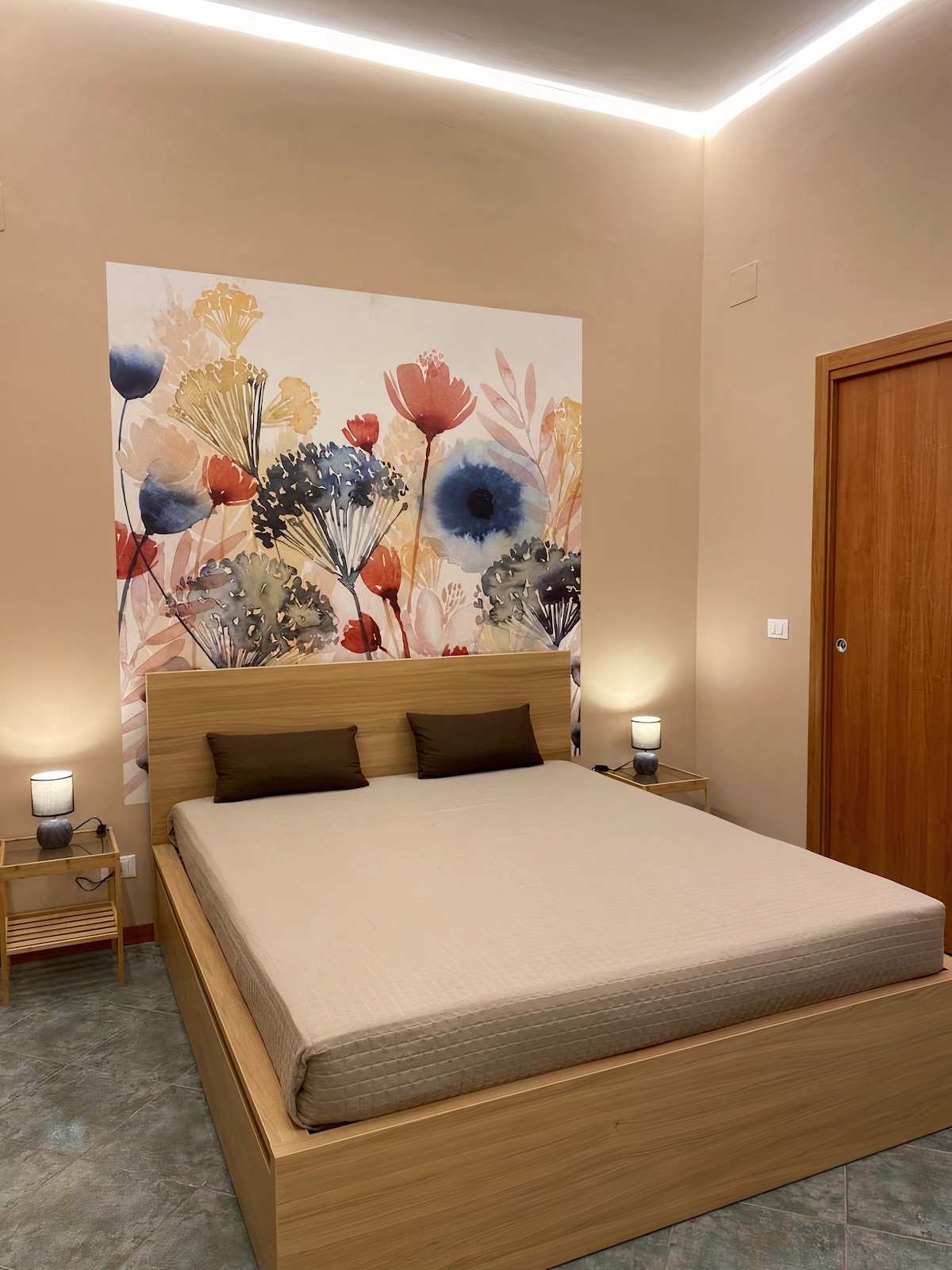 A serene bedroom is displayed, featuring a spacious bed with neutral bedding and two decorative pillows. On the wall, a floral mural adds color. Two bedside tables with lamps provide soft illumination, complementing the warm tones of the room.