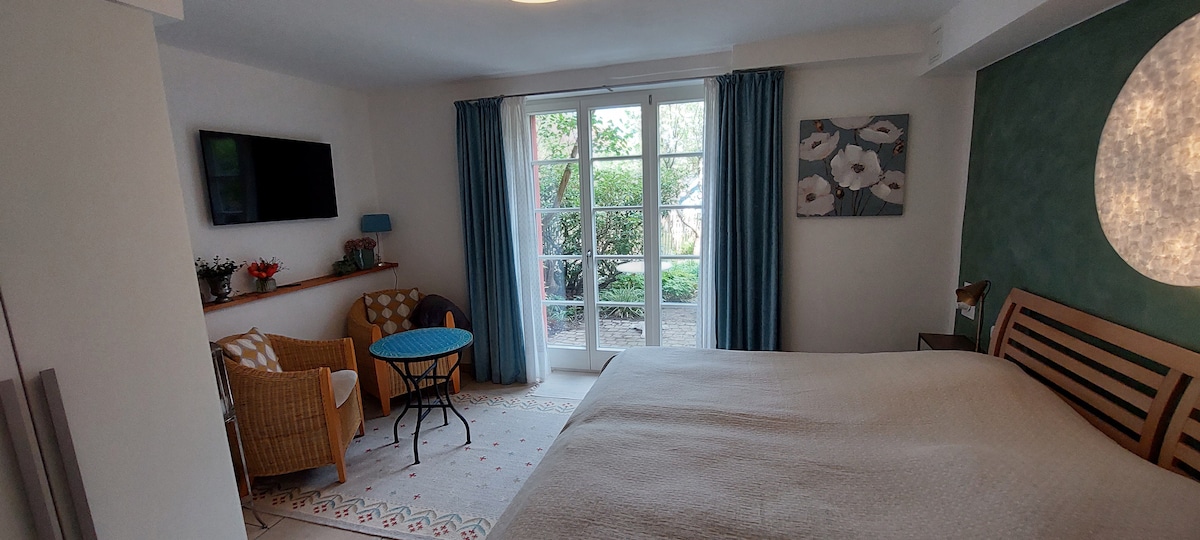 A bright bedroom features a spacious double bed and a seating area with two wicker chairs and a round table. Natural light enters through glass doors that open to a small terrace surrounded by greenery. Decorative elements include a wall-mounted TV and artwork.