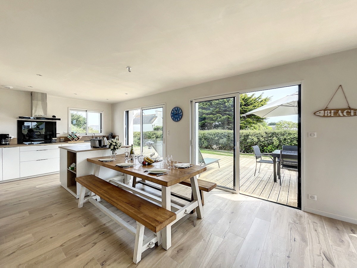 The dining area is highlighted by a large wooden table with benches, set for a meal. Large sliding doors provide access to the outdoor terrace, which features a shaded seating area and greenery visible beyond. The modern kitchen is equipped and open to the dining space.