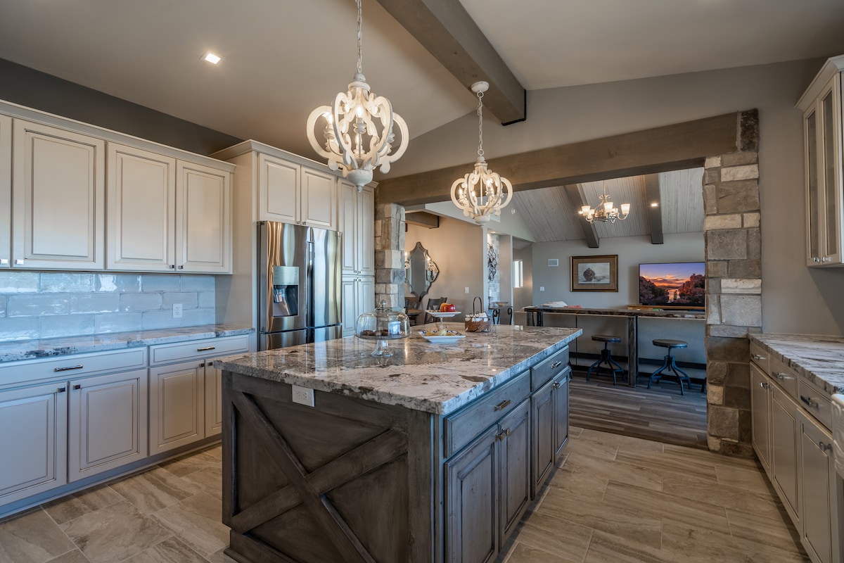A modern kitchen featuring light-colored cabinetry with sleek hardware and a spacious island with a textured countertop. Two decorative chandeliers hang above, illuminating the area. A doorway leads to an adjacent open space that includes seating and an entertainment area.