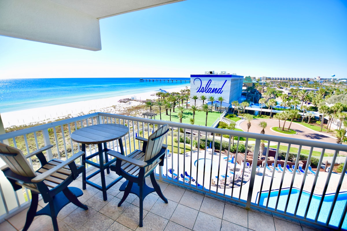 A spacious private balcony offers a scenic view of the beach and pier, complemented by a pool area below. Two outdoor chairs and a small table are positioned for relaxation, enhancing the outdoor experience with clear skies and serene ocean waters.