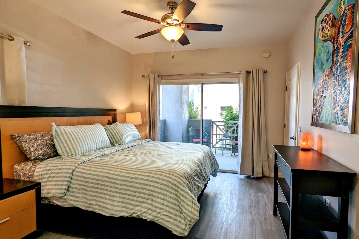 A large master bedroom features a spacious bed with striped bedding. Natural light is invited through sliding glass doors leading to a cozy patio, which is framed by light curtains. A ceiling fan and wooden furniture add to the room’s functionality.