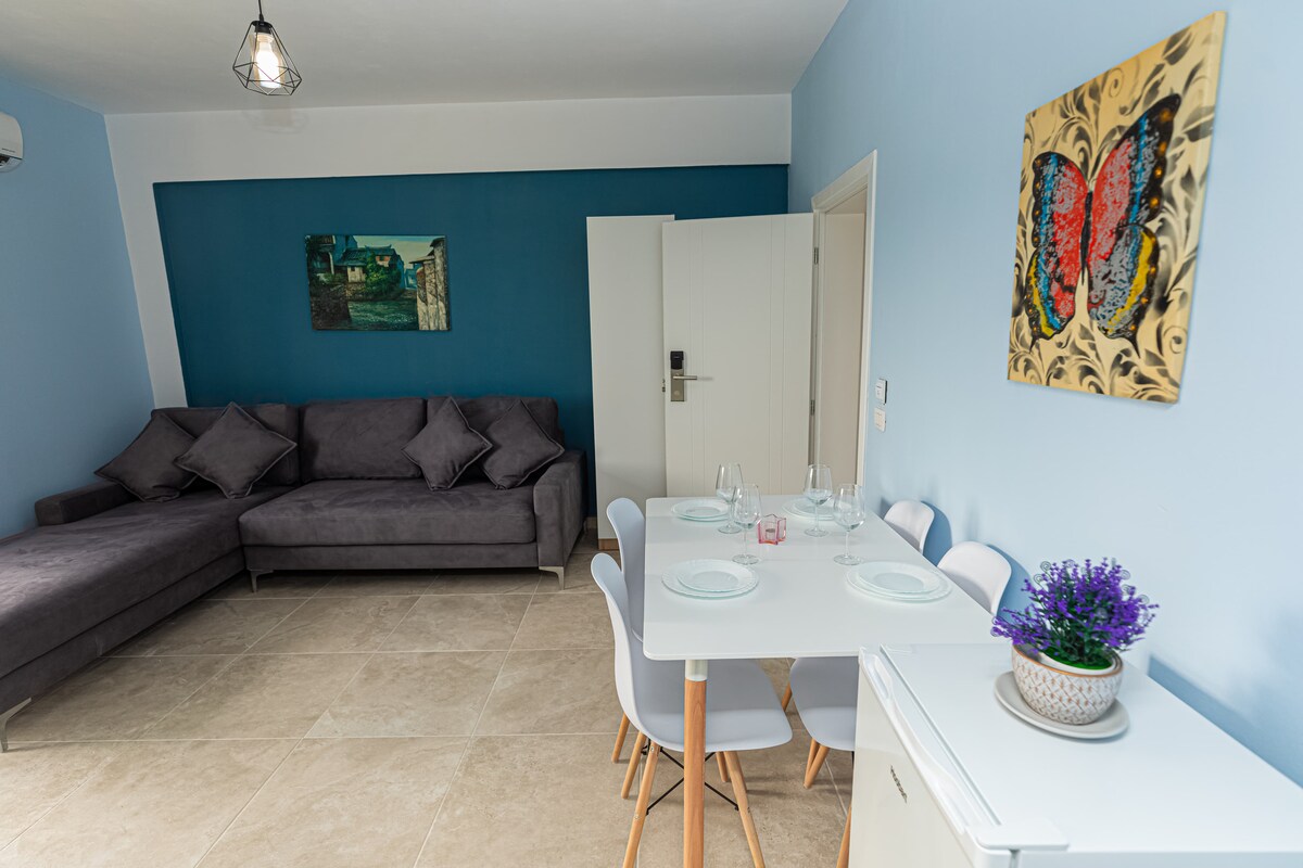 A modern living area features a gray sectional sofa adorned with plush cushions. A dining table with white chairs is set for a meal, accompanied by a decorative plant. Artwork on the walls adds a touch of color against the blue and white backdrop.