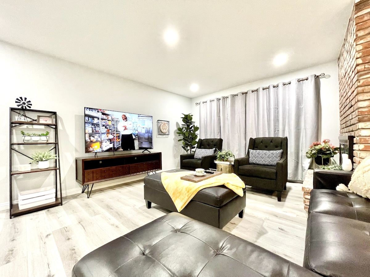 A modern living area features a large flat-screen TV mounted on the wall, with a cozy arrangement of two armchairs and a sectional sofa. Natural light filters through floor-length curtains, while decorative plants and a side table add a touch of greenery and sophistication to the space.