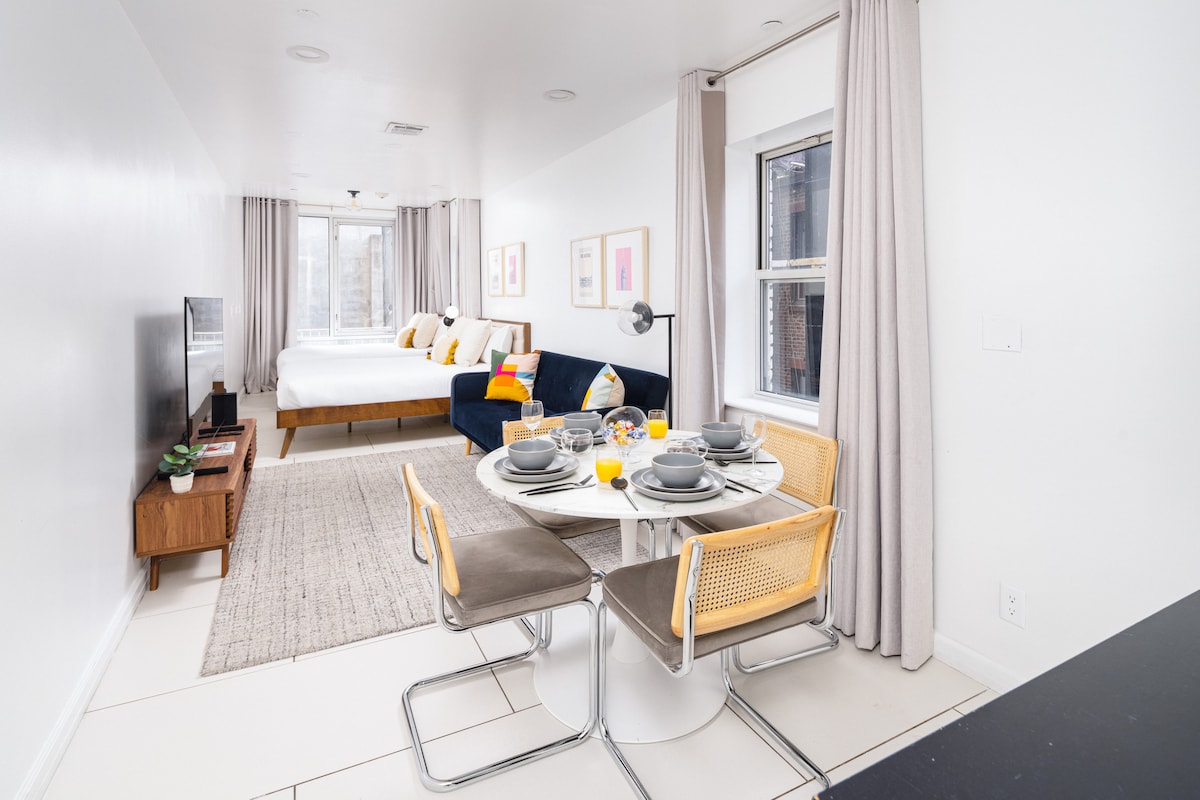 A modern studio layout is displayed, featuring a spacious sleeping area with a large bed and a pullout sofa. A round dining table is set for four, with tableware arranged neatly. Natural light floods in through large windows dressed with light curtains.