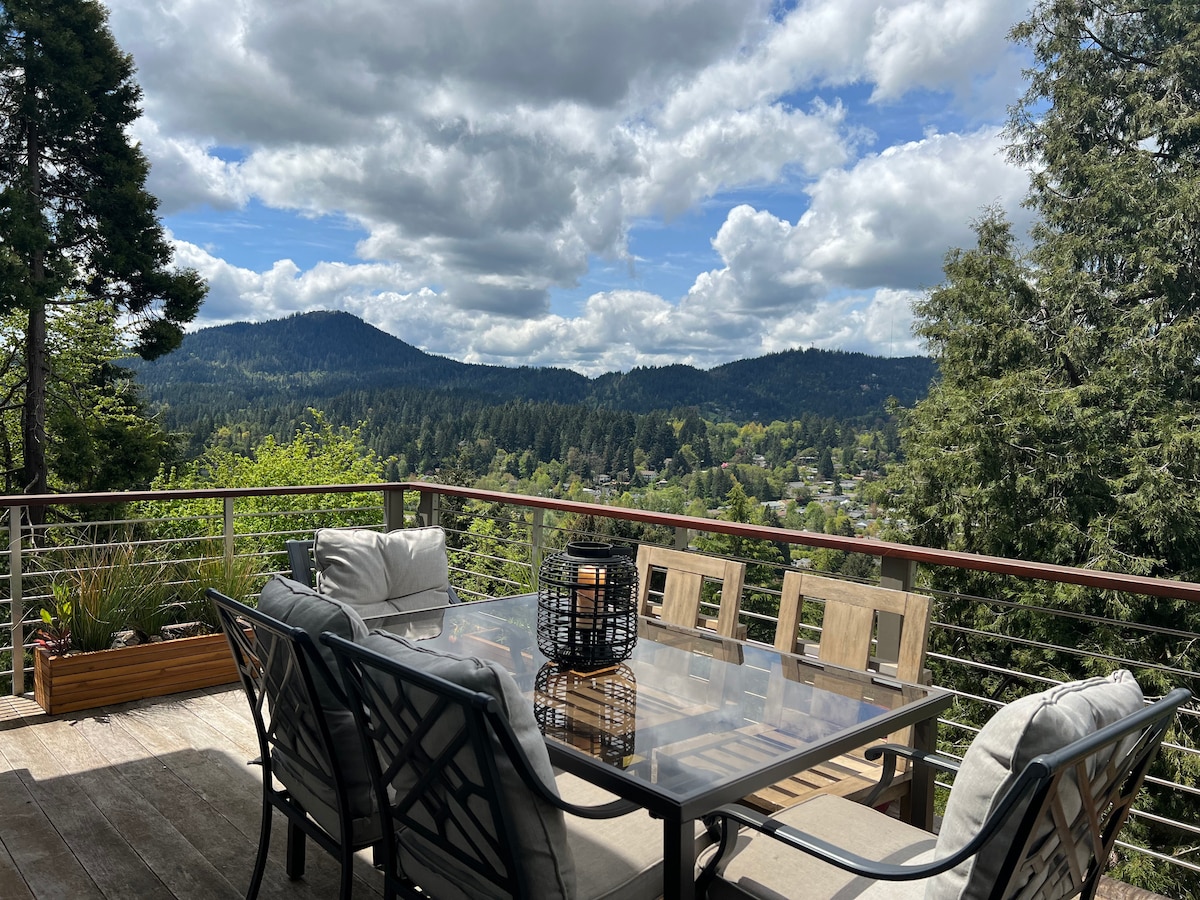 An outdoor patio area is presented with a glass-top dining table surrounded by comfortable chairs. Lush greenery and rolling hills create a scenic backdrop, while a lantern adds a touch of softness. Partly cloudy skies above enhance the tranquil setting.