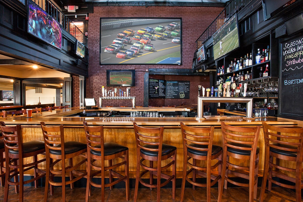 The spacious bar area features a long wooden counter lined with high-backed chairs. A large television is mounted on the brick wall, showcasing a racing event. Surrounding the bar, multiple screens are visible, displaying various sports events. The warm wood tones create a casual, inviting setting.