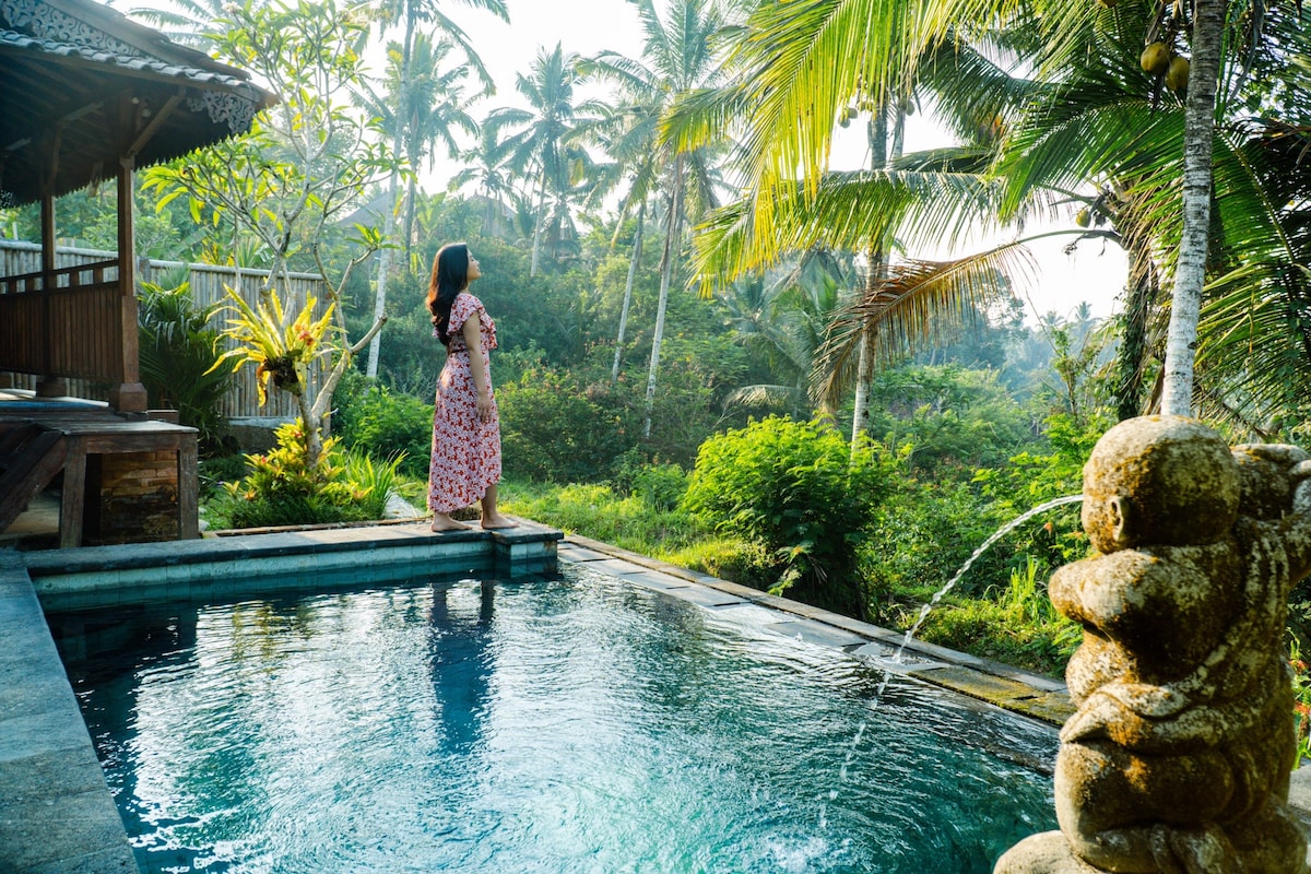 A tranquil private pool is surrounded by lush greenery and tropical palm trees. Natural mist softly blankets the area, and a gentle water feature adds to the serene atmosphere. A covered terrace with wooden details offers a relaxing view of the picturesque landscape.