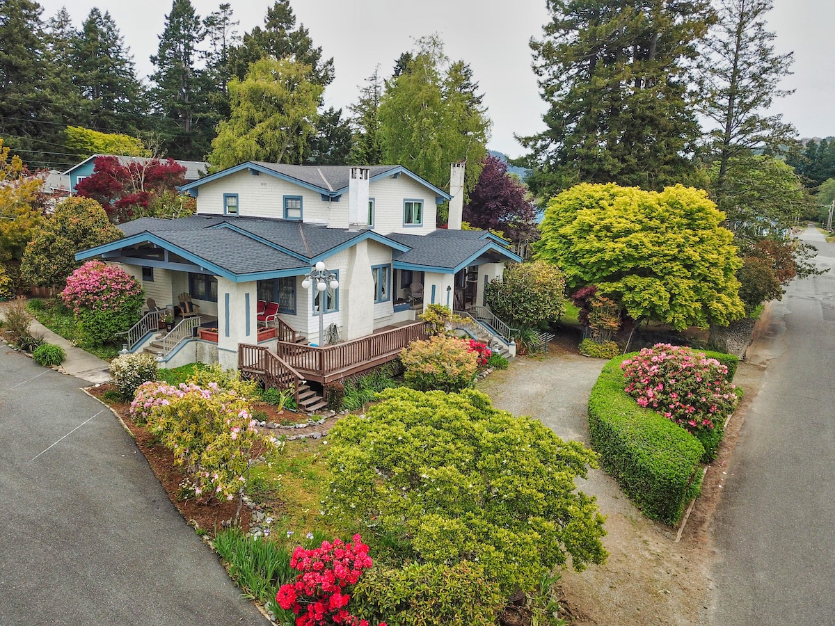 A spacious residential property is surrounded by lush greenery and vibrant flower beds. A welcoming porch is accessible by a wooden walkway, and large trees provide ample shade. The setting creates a serene atmosphere, ideal for relaxation and privacy.