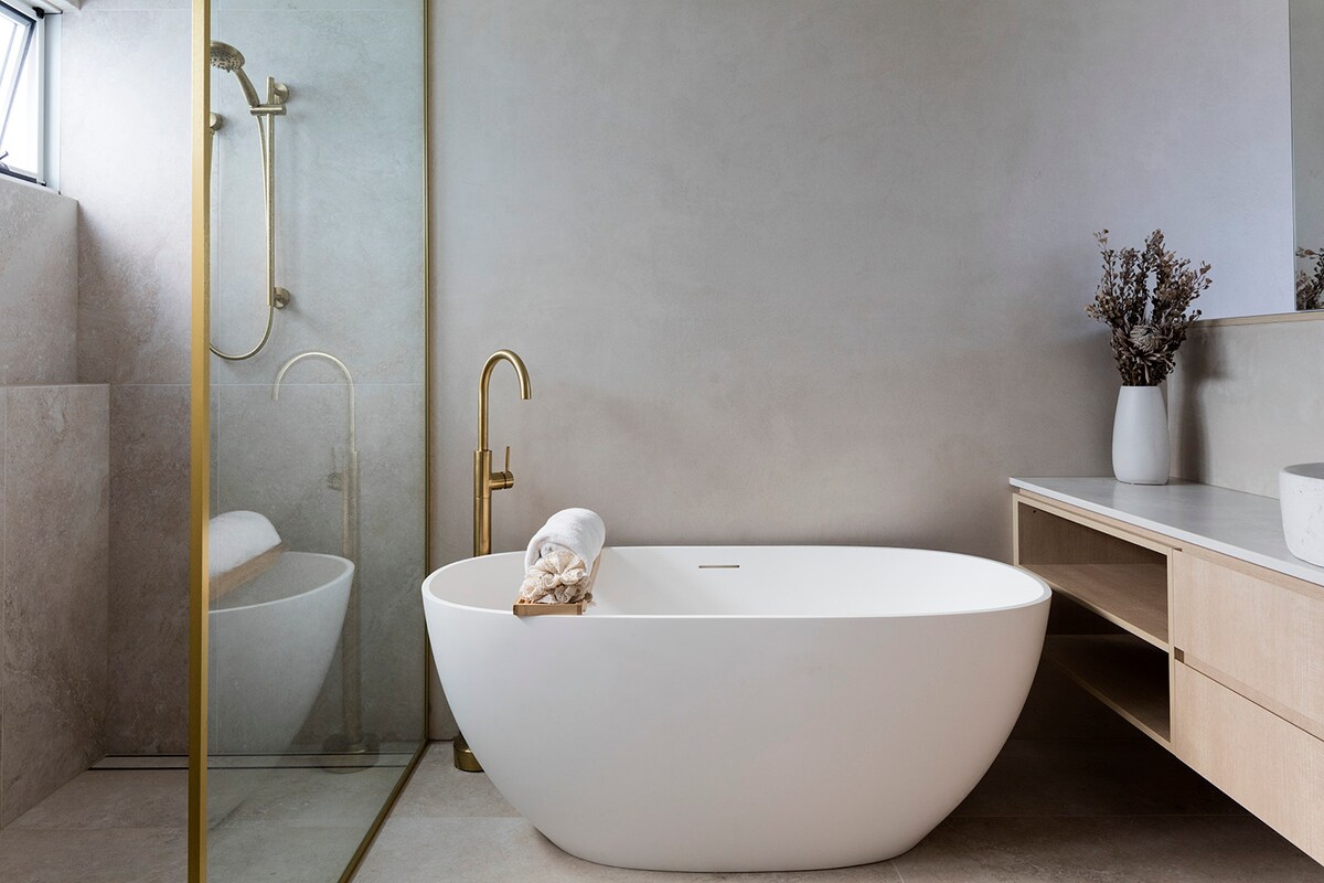 A modern bathroom features a freestanding white bathtub with a towel neatly placed on the edge. A glass shower enclosure reflects natural light, while soft beige walls and a minimalist wooden vanity create a serene environment. A decorative vase adds a touch of elegance.