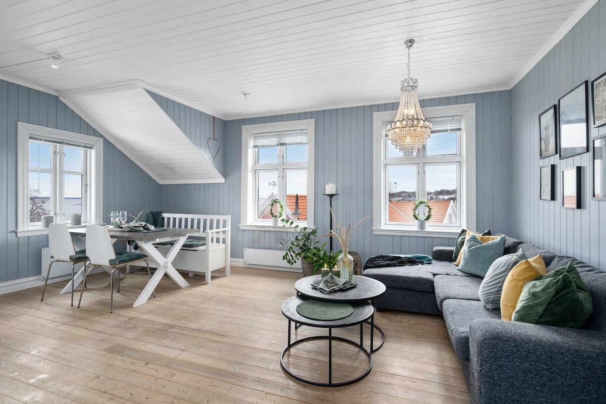 A light-filled living area features a spacious sectional sofa adorned with vibrant throw pillows. A contemporary chandelier hangs from the ceiling. A dining table with four chairs is positioned near large windows, offering views and natural light. Wooden flooring complements the overall design.