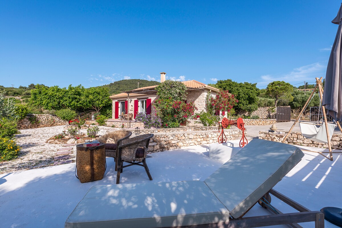 A serene outdoor area showcases a sunbed positioned on a light-colored surface, with a backdrop of a stone house featuring red shutters. Lush greenery and colorful flowering plants surround the space, providing an inviting environment for relaxation.
