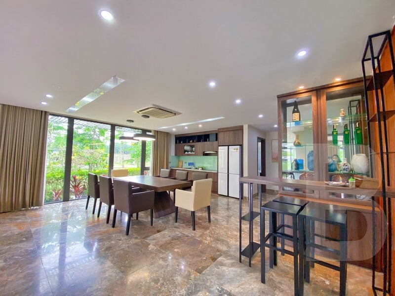 A spacious living and dining area features large windows that provide natural light and a view of the lush outdoor landscape. A modern dining table is surrounded by six chairs, while a stylish kitchen area includes sleek cabinetry and essential appliances, seamlessly integrated into the space.