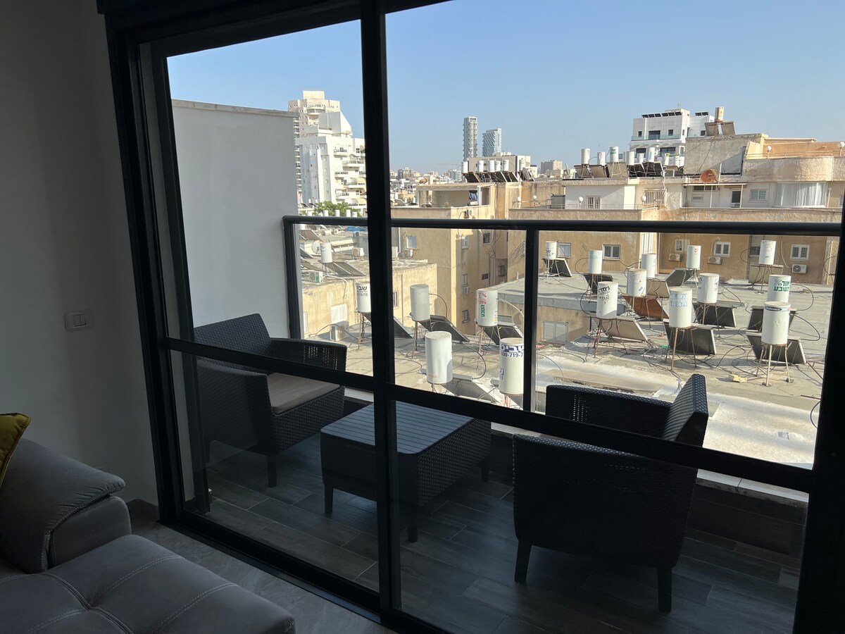 A balcony area is visible, featuring two black wicker chairs and a small table. The surrounding view includes various rooftops and air conditioning units, with a clear blue sky above, creating a sense of openness.