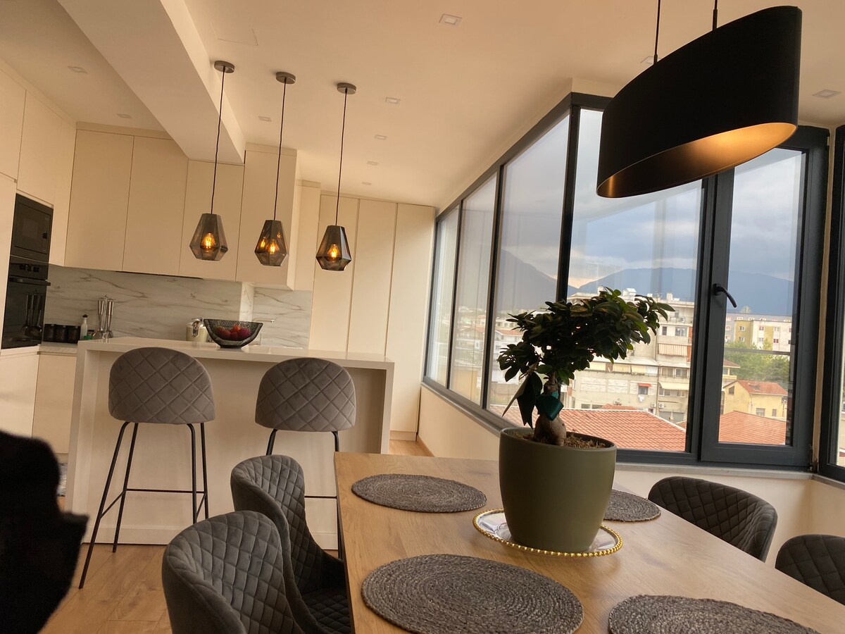 A modern kitchen and dining area feature large windows offering mountain views. A round dining table with woven placemats is surrounded by textured chairs. Pendant lights hang above, and a potted plant sits at the center of the table, enhancing the minimalist design.