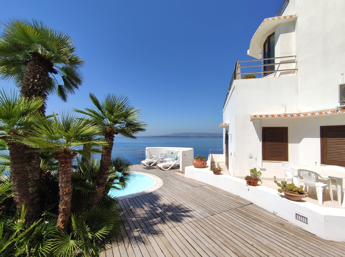 An outdoor area features a wooden deck surrounded by lush greenery and palm trees. A small infinity pool reflects the clear blue sky, while a seating area with white chairs and a table offers views of the tranquil sea.