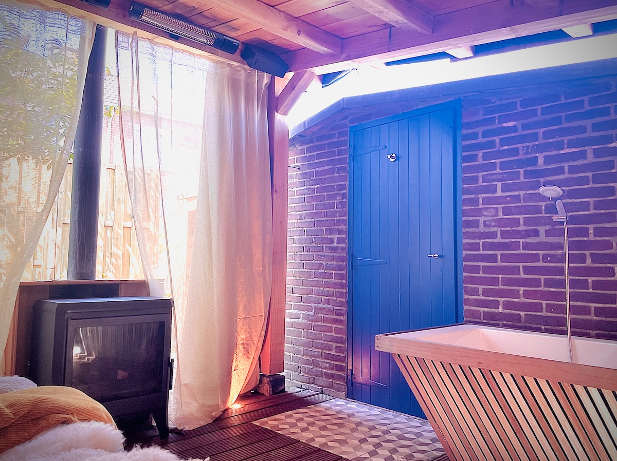 An inviting interior space features a modern bathtub positioned next to a small fireplace. Sheer curtains softly filter natural light, enhancing the warm atmosphere. A vivid blue door is seen in the background, with textured brick walls providing a rustic touch.