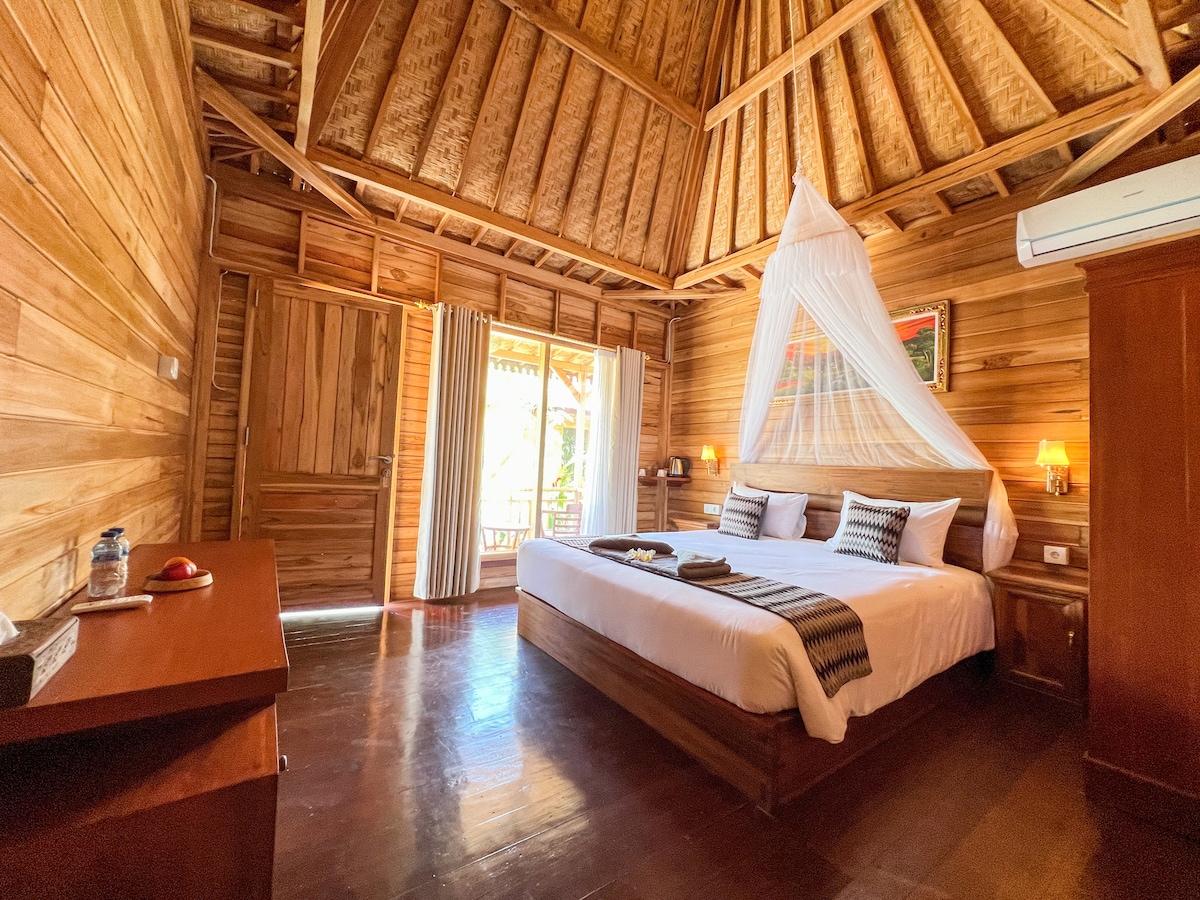 A spacious bedroom features wooden walls and a high ceiling with exposed beams. A large bed is dressed with neutral linens and a decorative throw. Soft lighting is provided by lamps on either side, and large windows allow natural light to fill the room.