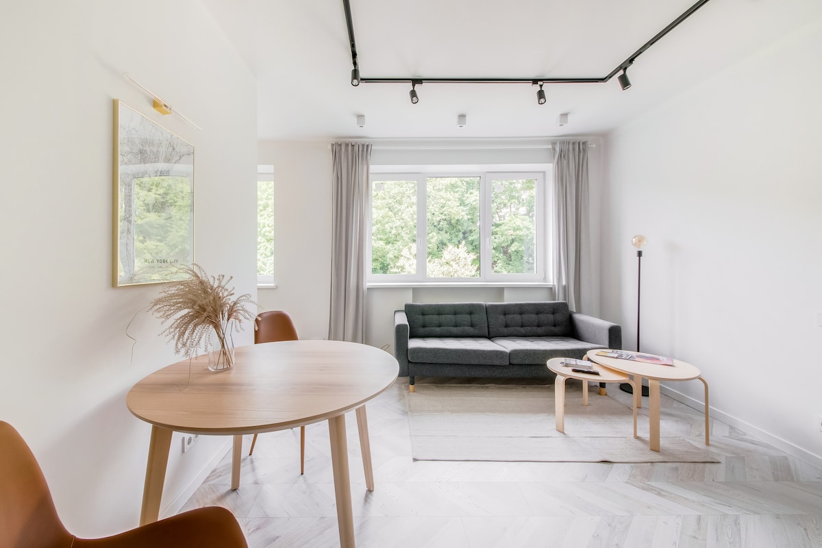 A spacious living area features a dark grey sofa centered against a light wall, with two wooden coffee tables placed in front. Natural light enters through wide windows adorned with sheer curtains, and a round dining table is set to the left, accompanied by modern chairs.