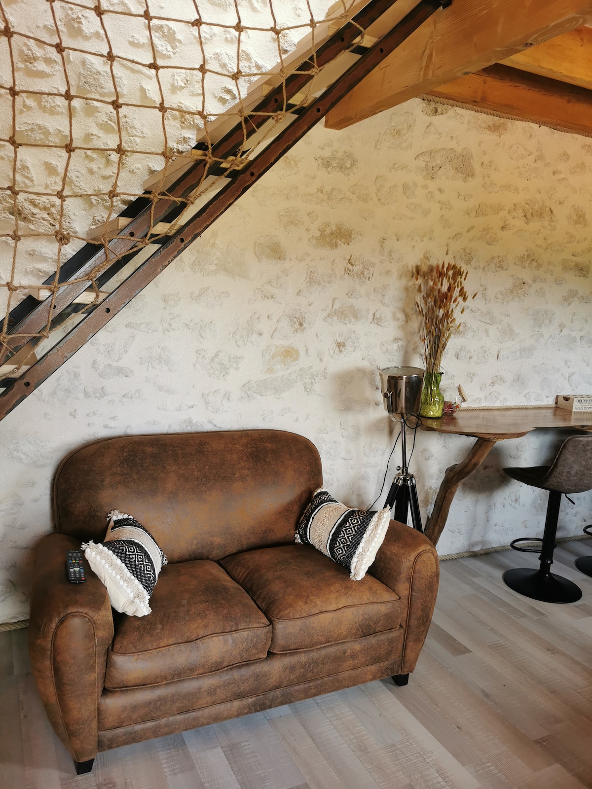 A cozy sitting area features a brown upholstered loveseat adorned with decorative cushions. A rustic wooden table and modern chairs accompany the space, while a decorative vase filled with dried flowers adds a touch of natural elegance. A staircase with a unique railing leads to the upper level.