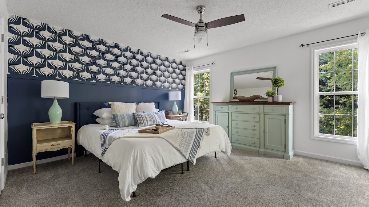 A serene bedroom is presented with a spacious bed dressed in soft white linens. Decorative blue wallpaper adds a subtle pattern, while natural light enters through two windows. A light green dresser and a bedside table with a lamp complete the inviting space.