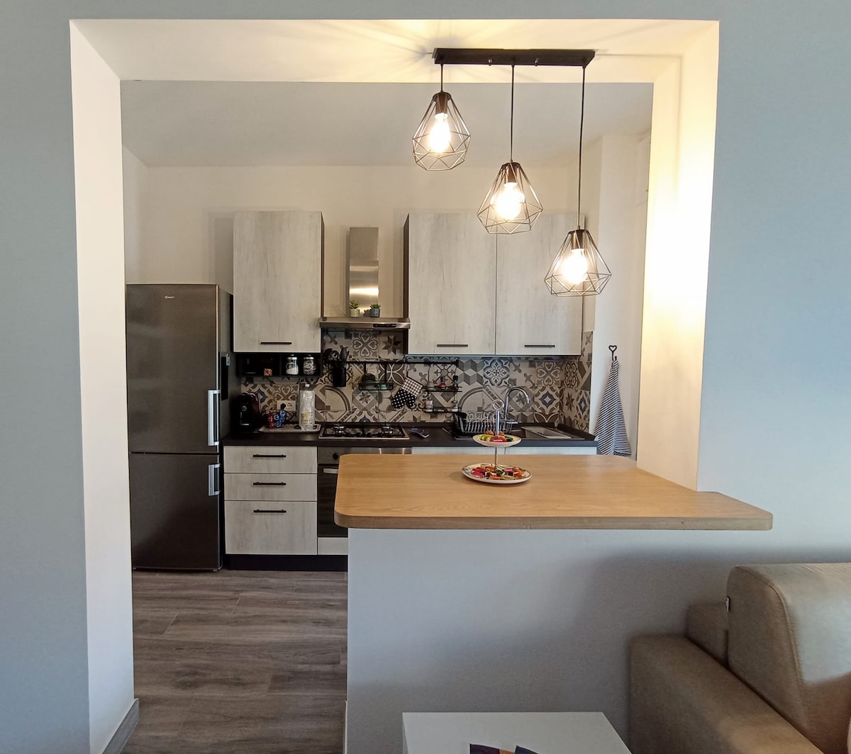 The kitchen area features modern cabinetry and appliances, illuminated by three pendant lights. A clear countertop displays a plate of colorful fruit, while a stainless steel refrigerator stands nearby. Decorative tile backsplashes add a subtle texture to the space.