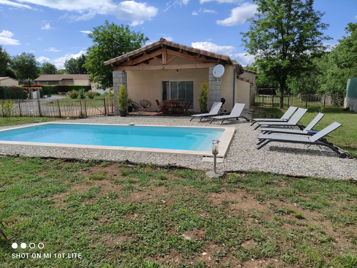A private outdoor area features a rectangular swimming pool surrounded by a gravel patio. Four lounge chairs are positioned beside the pool, and a wooden dining table with chairs is visible under a shelter. Lush greenery and trees frame the space.