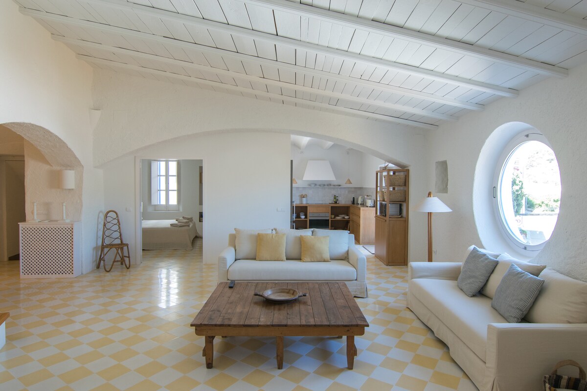 A spacious living area features a light color palette with high, beamed ceilings and large round windows. A wooden coffee table sits centrally, surrounded by a comfortable sofa. The adjacent kitchen area is visible in the background, enhancing the open-plan layout.