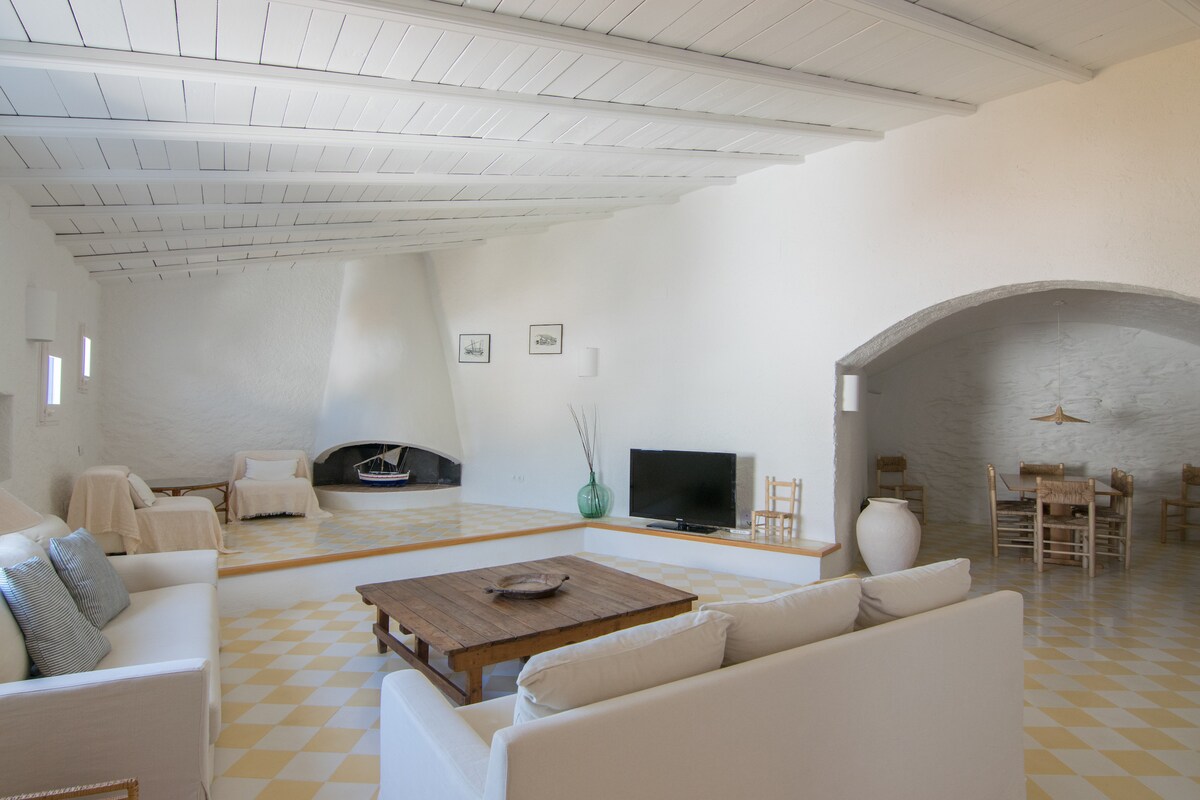 A spacious living area is featured, with light-colored walls and a high, beamed ceiling. A cozy seating arrangement surrounds a wooden coffee table, while a minimalist television set stands nearby. An dining space integrates into the room, with a round table and simple chairs visible.