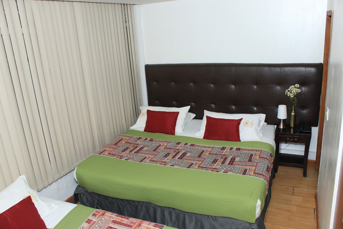 The room features two spacious beds, each adorned with decorative pillows in white and red. A patterned throw blanket is draped across the larger bed. Soft natural light filters through vertical blinds, illuminating the room's warm wood flooring and simple bedside table.