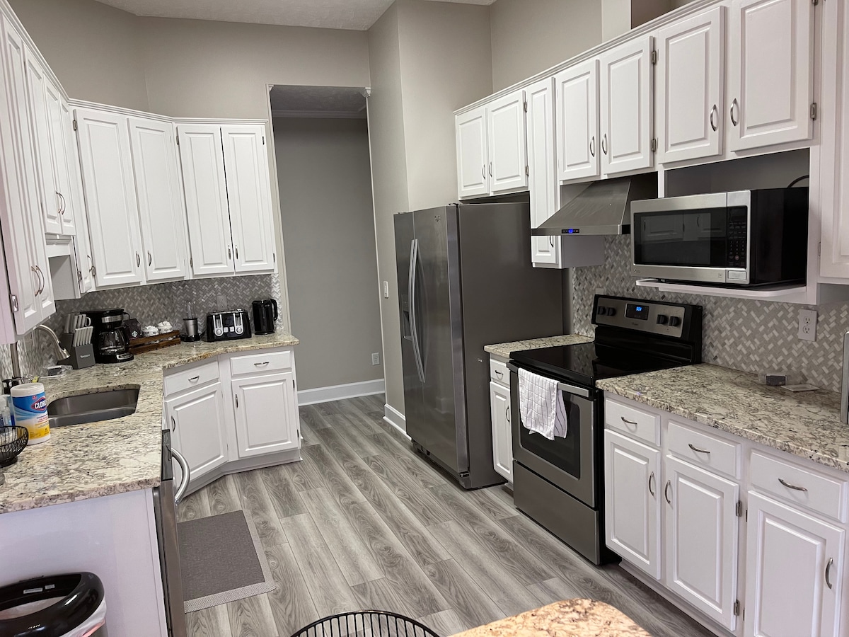 The spacious kitchen features white cabinetry and gray granite countertops, creating a bright and modern environment. Stainless steel appliances, including a refrigerator and oven, are visible, along with a coffee maker and toaster on the counter. The layout offers ample storage and workspace.