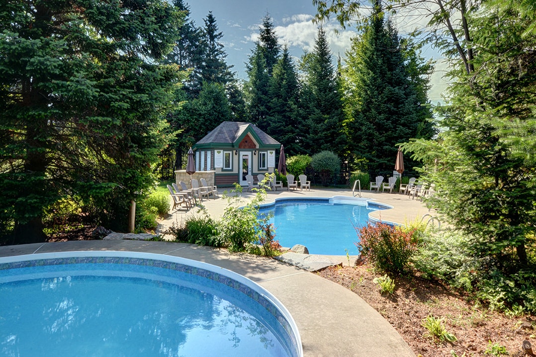 Two shared swimming pools are surrounded by lush greenery and tall trees, providing a serene setting. A nearby gazebo offers seating and shade, making it an inviting space for relaxation. Several lounge chairs are positioned around the pools, enhancing the tranquil atmosphere.