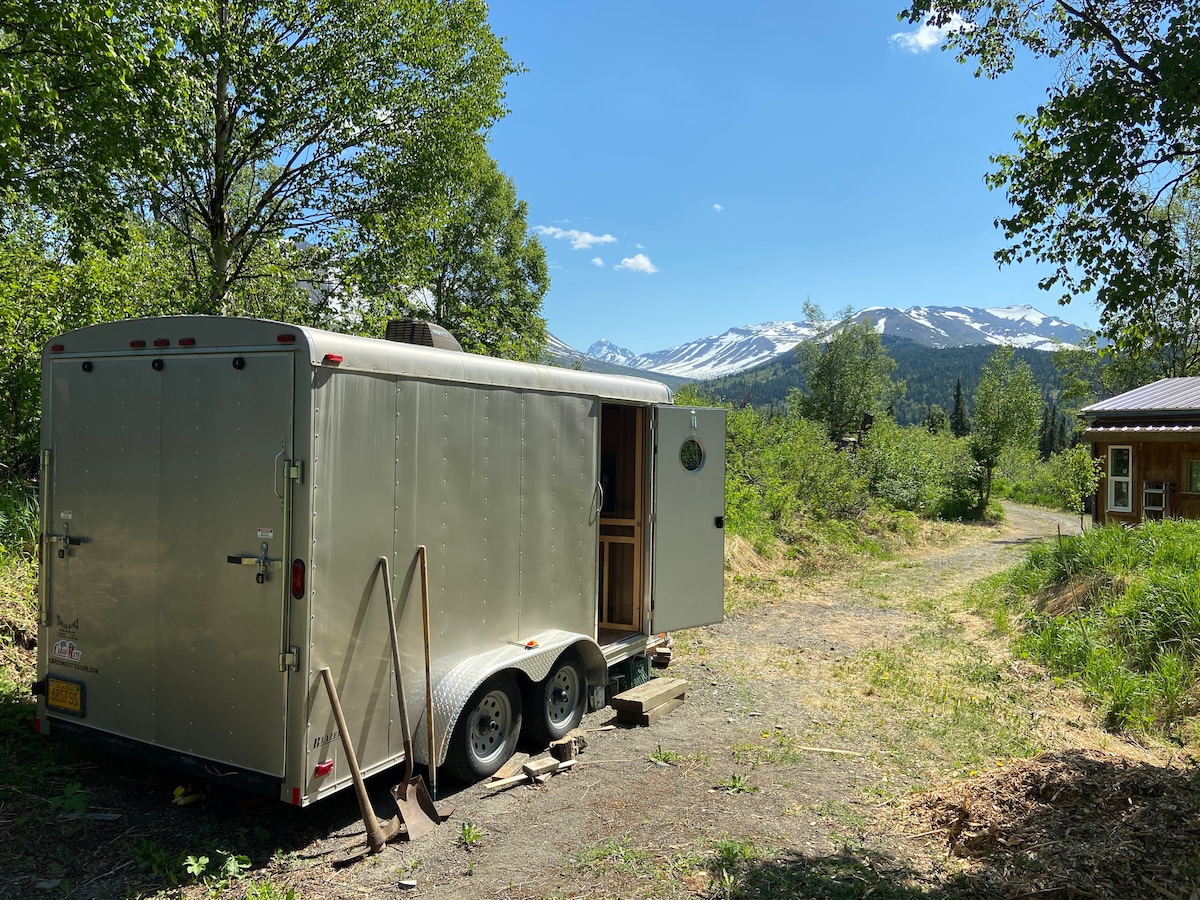 A converted trailer is positioned on a forested lot, showcasing a gravel path leading to a distant cabin. Snow-capped mountains and clear blue skies provide a scenic backdrop, while trees surround the area, emphasizing the natural beauty and tranquility of the location.