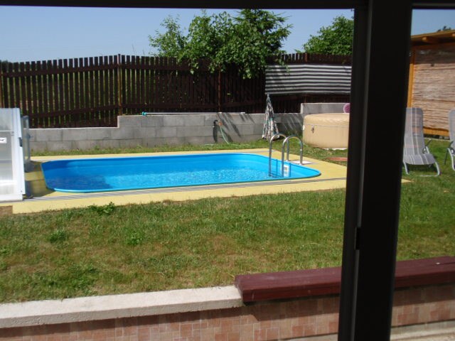 A private outdoor area features a rectangular swimming pool surrounded by a grassy lawn. Sun loungers are positioned nearby, and a hot tub is visible to the right. A wooden fence provides privacy, while trees and shrubs add greenery to the space.