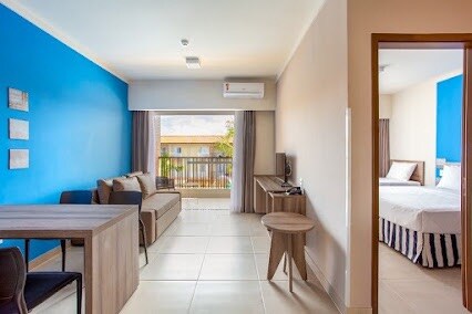 A bright living area features a blue accent wall and neutral-toned furnishings, including a dining table and a sofa. A smart TV is mounted on the wall, and large windows provide natural light. The entrance to a separate sleeping area is visible in the background.