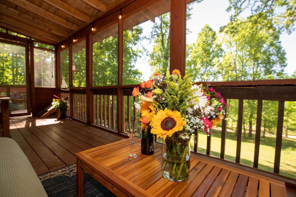 A cozy screened-in deck is highlighted by a wooden table featuring a vibrant bouquet of flowers, including sunflowers. The inviting space is surrounded by trees, offering a connection to nature. Soft light filters through the screens, enhancing the relaxed atmosphere.