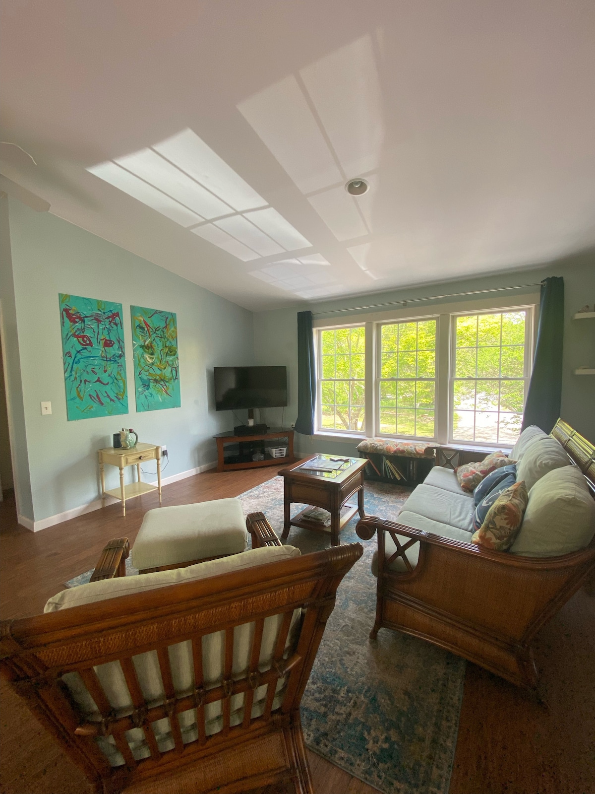 A bright living room features large windows allowing natural light to fill the space. Two colorful wall art pieces adorn the light blue walls. A comfortable seating arrangement includes a white sofa and wicker chairs set around a coffee table, creating an inviting atmosphere.