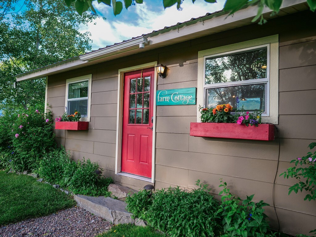 The exterior of the Farm Cottage features a welcoming red door and flower boxes filled with vibrant blooms. Lush greenery surrounds the cottage, enhancing its charm. The structure is modest in size, with neatly placed stones leading up to the entrance.