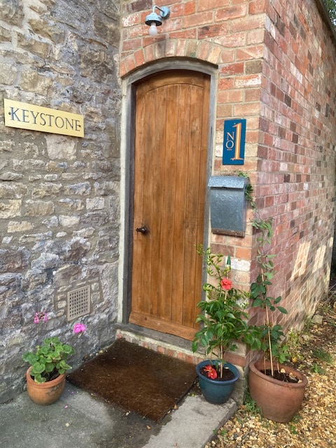 A charming entrance to Keystone is framed by weathered stone and warm brickwork. A solid wooden door with a round handle leads into the home, complemented by potted flowers and greenery. An engraved plaque indicates the house number, adding a personal touch.
