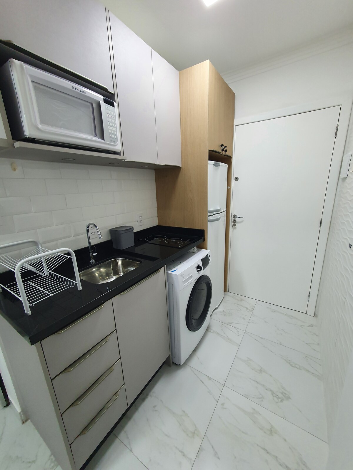 A compact kitchen area features modern cabinetry in neutral tones. A microwave, sink, and stovetop are conveniently arranged on a dark countertop. A washing machine is included, and a refrigerator stands to the side. Soft white tiles add to the clean, functional design.