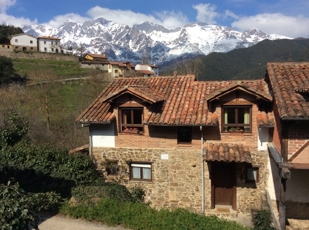 A rustic house features stone and wooden elements, with a tile roof and green windows. In the background, snow-capped mountains dominate the landscape, creating a serene natural setting. Nearby homes are subtly integrated into the hillside, enhancing the tranquil ambiance.