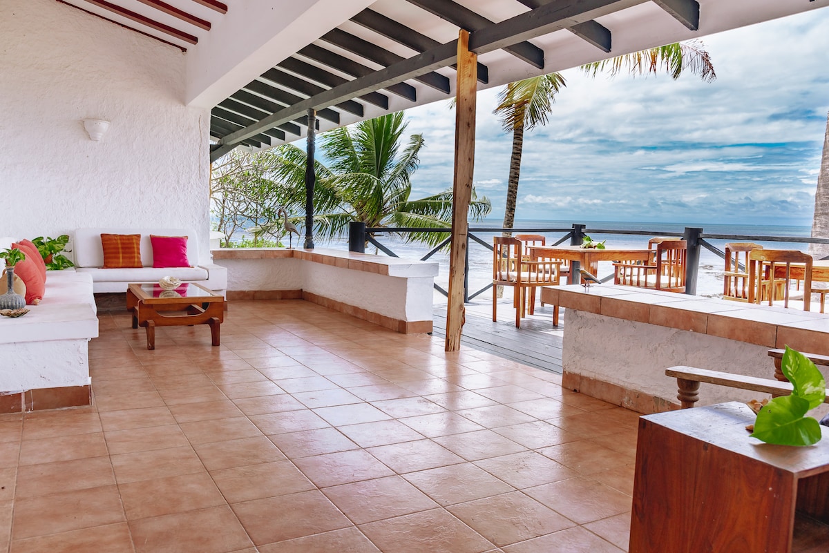 An open-air seating area features a white couch adorned with colorful cushions. A wooden table stands in the center, surrounded by windows. Tropical plants provide greenery, and views of the beach and ocean are visible in the background. The tiled flooring enhances the spacious feel.