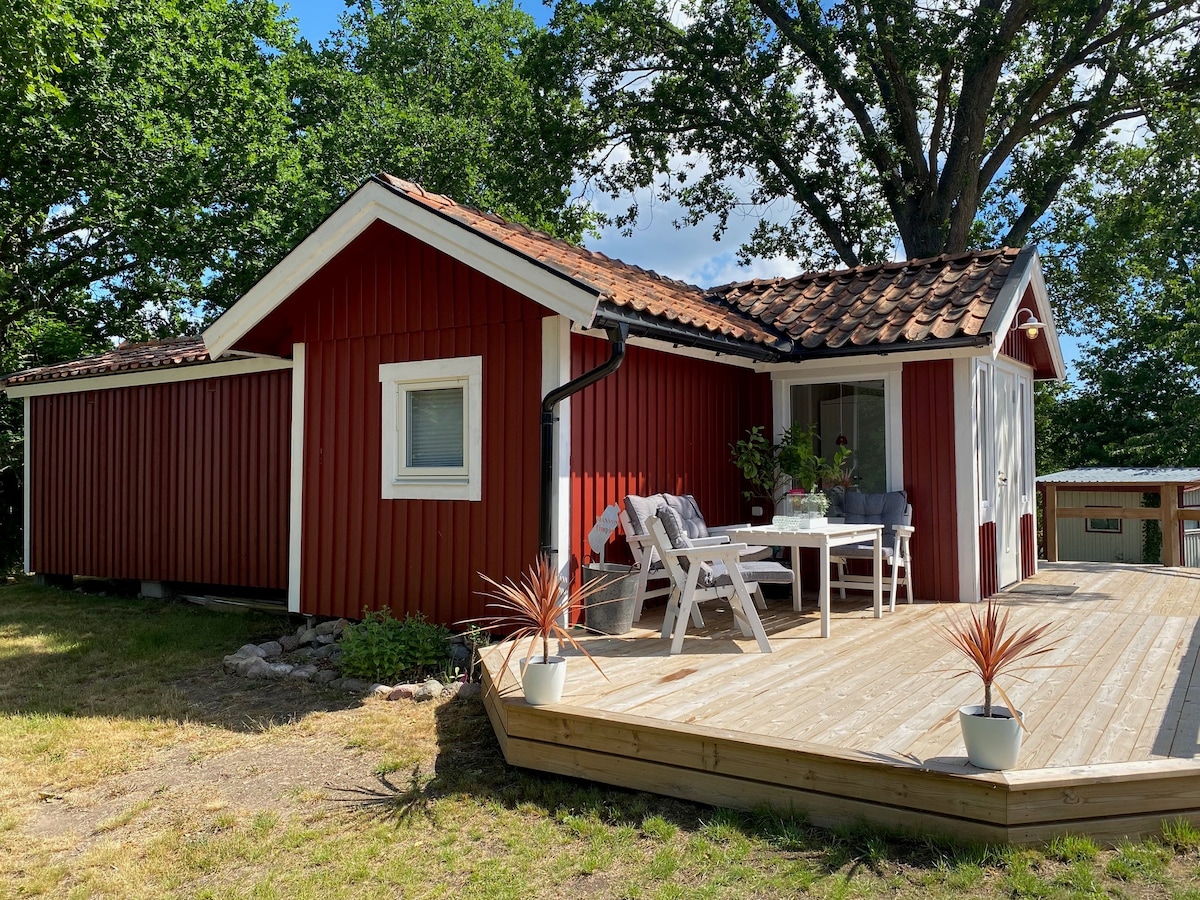 A charming red wooden cottage is positioned under a leafy oak tree. The front deck is adorned with a table and chairs, inviting relaxation. Large windows allow natural light to fill the interior, while planters with greenery offer a touch of nature.